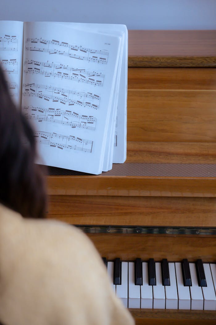 Back view of crop anonymous female in casual outfit playing on wooden piano with music book in light room